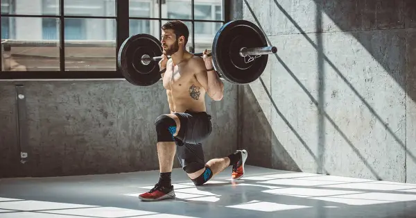 Shirtless athlete gym mein heavy barbell ke saath lunges karta, knee support pehle focus on form