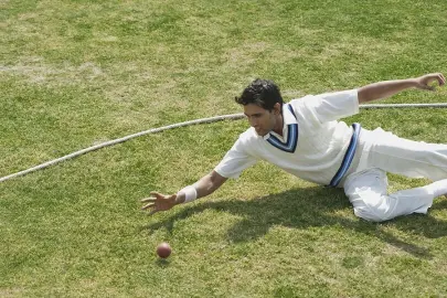 Player grass field pe slide karke low catch pakadta, white uniform mein pro fielding